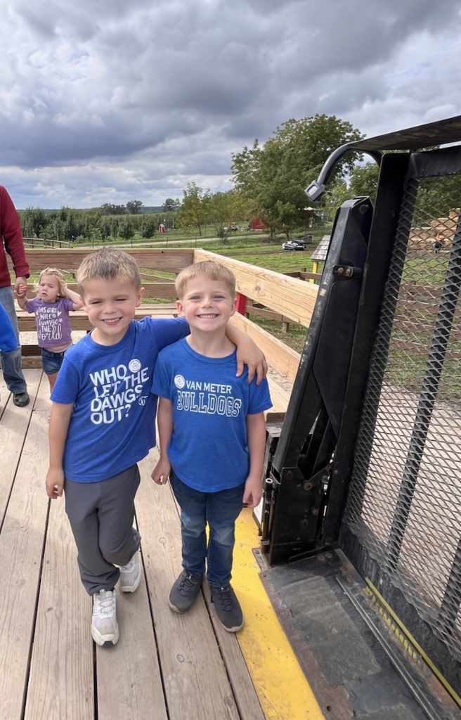 Two young boys smiling and posing together on a wooden deck with a farm field and cloudy sky in the background.