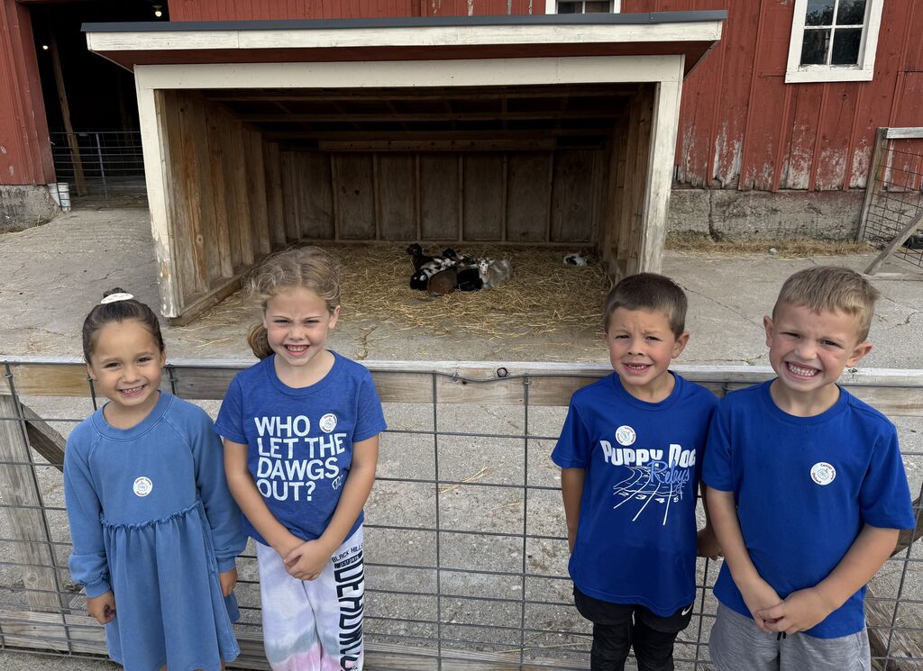 Four smiling children stand in front of a barn enclosure with a small calf resting inside on straw bedding.