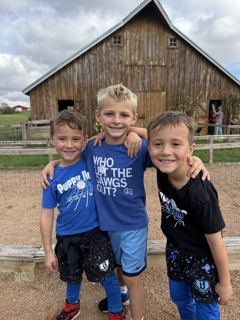 Three young boys smiling and posing with arms around each other in front of a rustic wooden barn on a cloudy day.