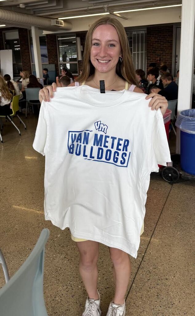 Smiling young woman holding up a white Van Meter Bulldogs t-shirt in a busy indoor setting.