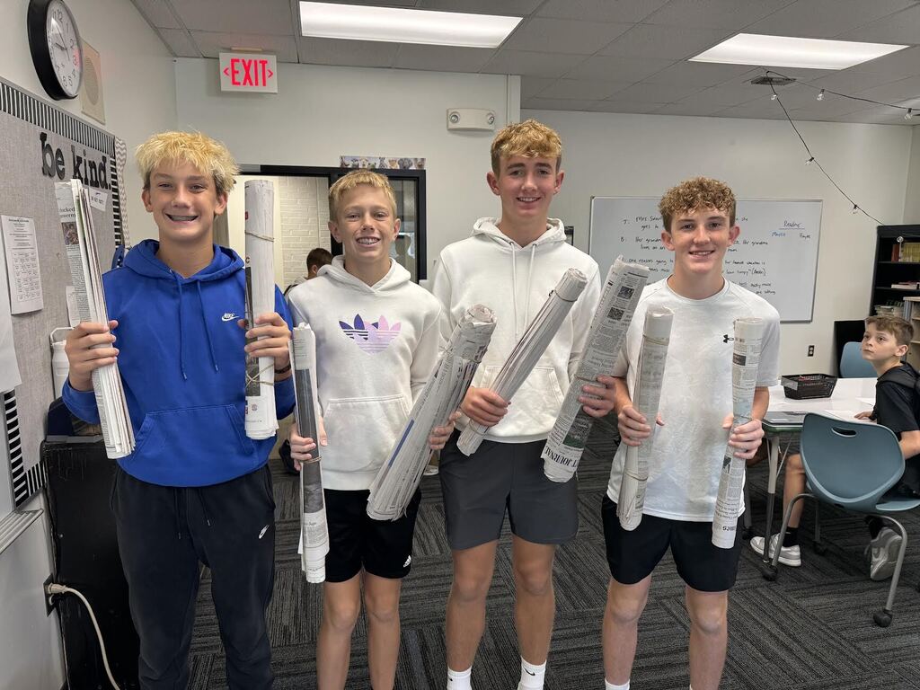 Four teenage boys standing in a classroom holding rolled-up posters, smiling at the camera.