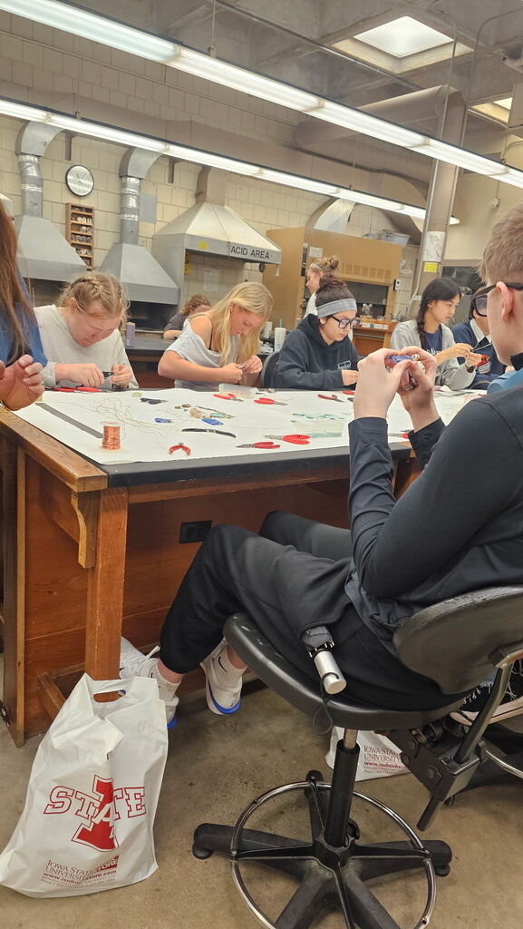 Students use pliers, wire, and small components to create jewelry around a large workbench in a studio.