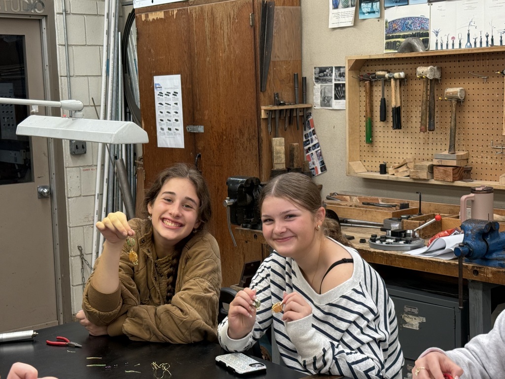 Two smiling students display handmade jewelry pieces at a workbench surrounded by studio tools.