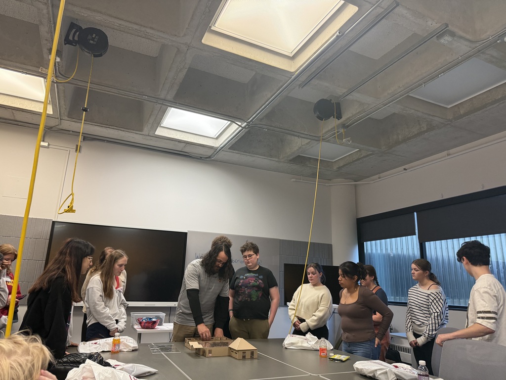 An instructor demonstrates a model to students gathered around a large worktable in a design classroom.