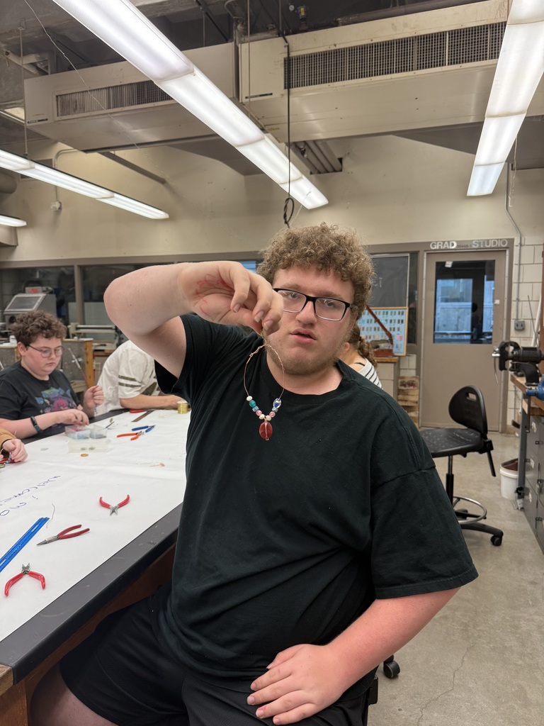 A student holds up a colorful beaded necklace while seated at a jewelry-making workbench.