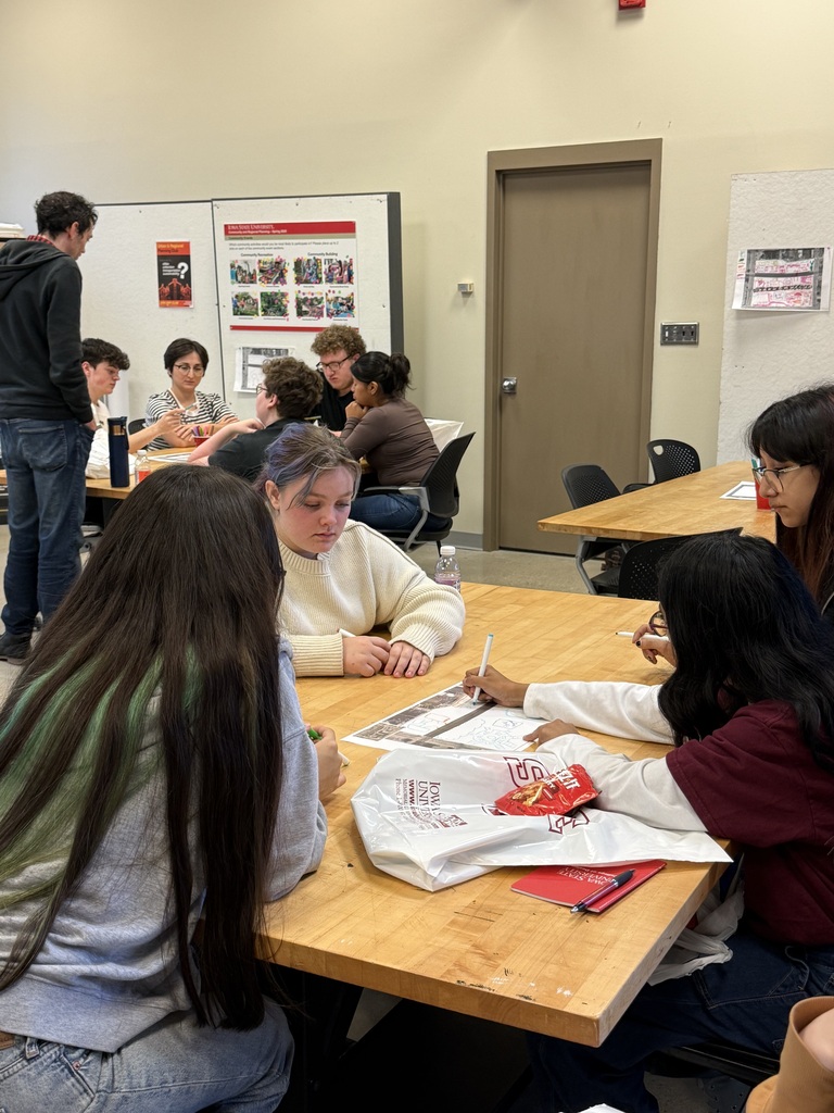 Students gather around classroom tables, discussing and drawing design ideas on printed worksheets.