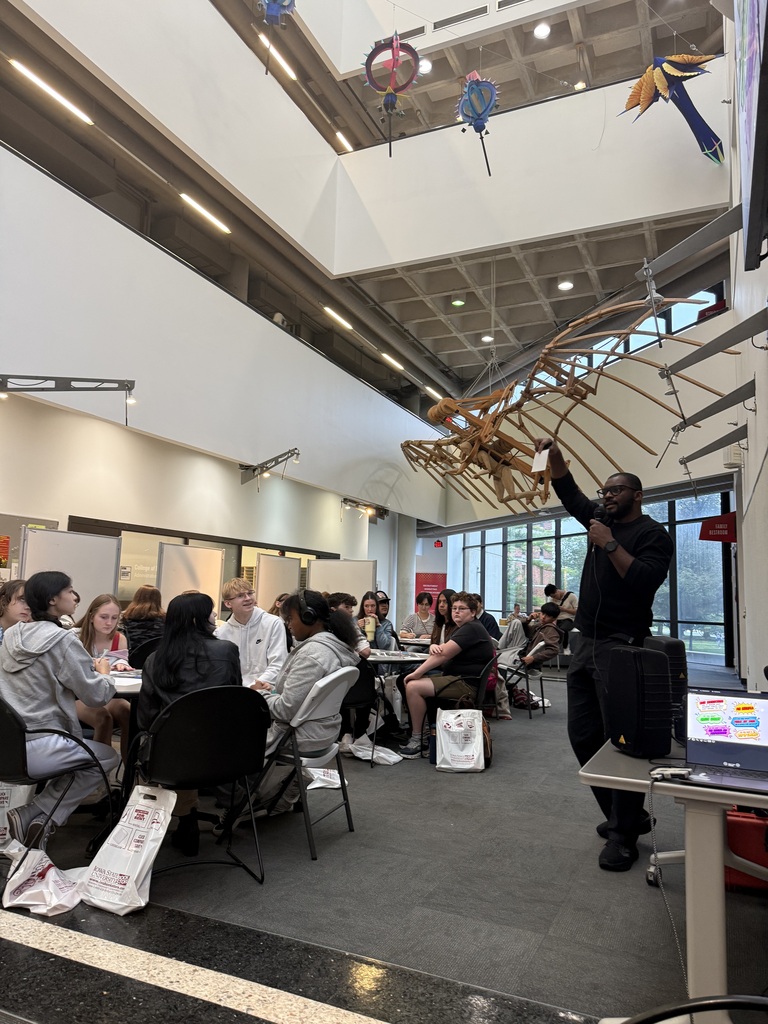A man stands presenting in front of a seated group inside a spacious room with a large wooden skeleton hanging overhead.