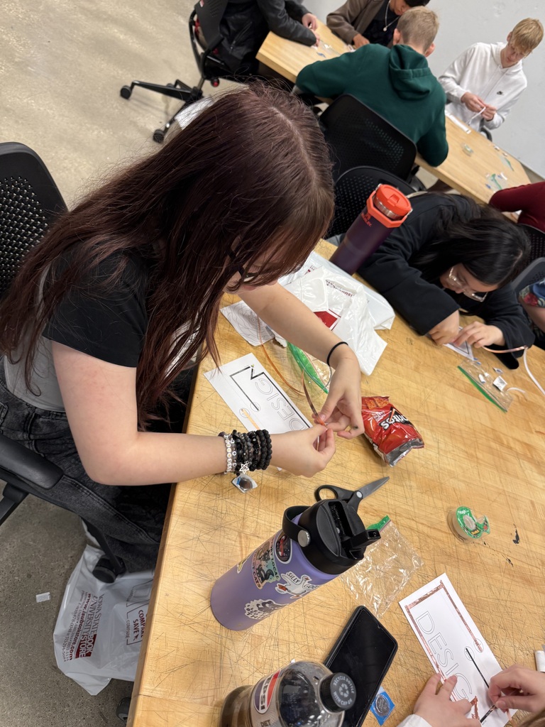 Students work with small craft materials, tubing, scissors, and design worksheets at a classroom table.