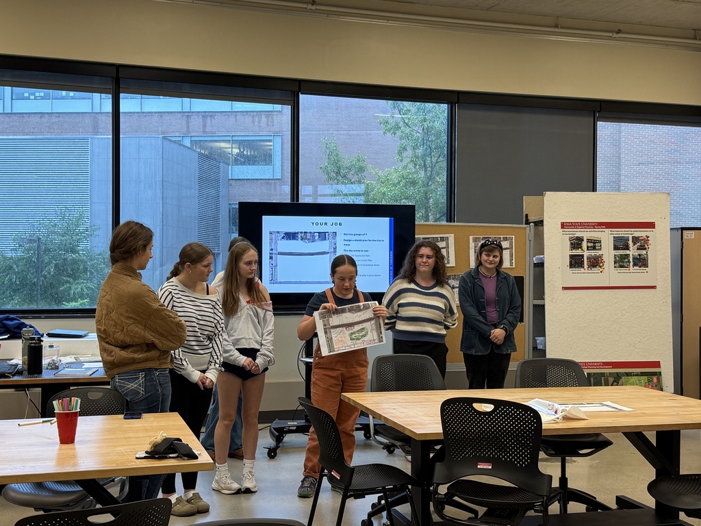Students present a design plan to classmates in a university classroom, holding a drawing beside a presentation screen.
