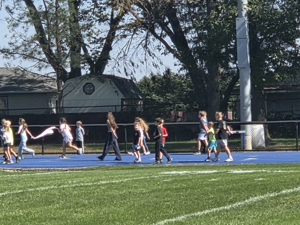 Children walking in a line along a blue track beside a grassy field under large trees on a sunny day.