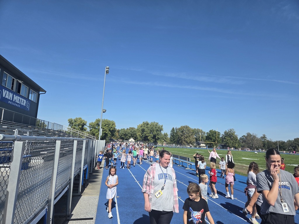 Crowd of children and adults walking on a blue track beside a sports field under clear skies.