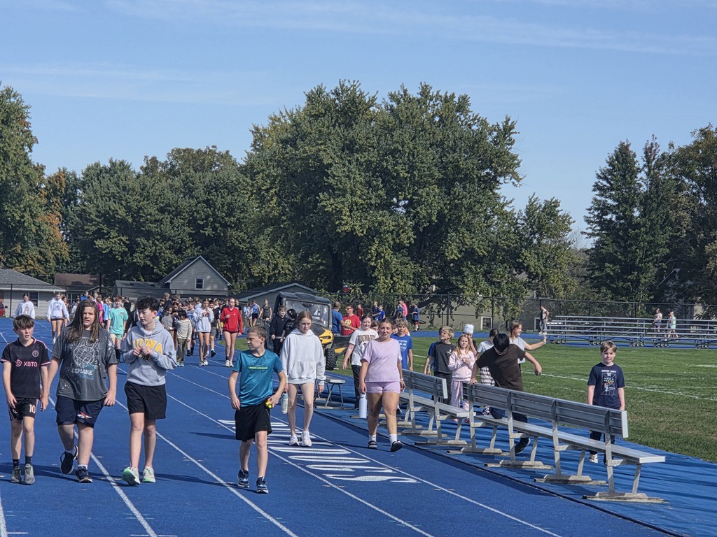 Group of students walking on a blue outdoor track near bleachers and green field under a clear sky.