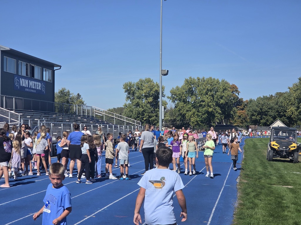 Children and adults gather on a blue running track near bleachers under a clear sky at Van Meter school stadium.