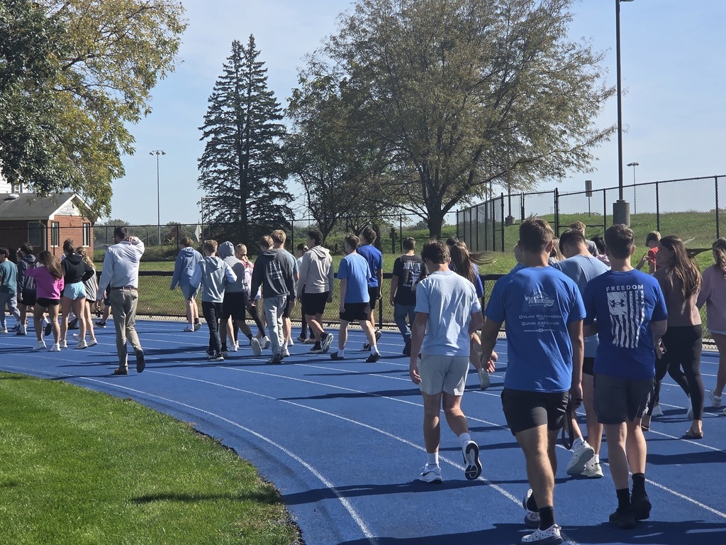 Group of people walking on a blue outdoor running track surrounded by trees on a sunny day.