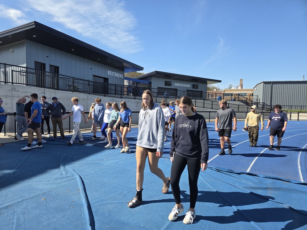 Group of young people walking on a blue outdoor sports court under clear skies near modern buildings.