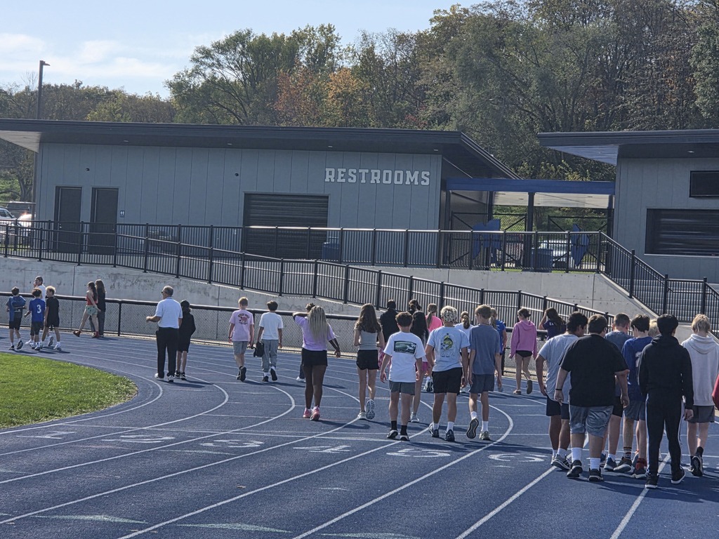 Group of students walking on a blue outdoor track near a building labeled RESTROOMS on a sunny day.