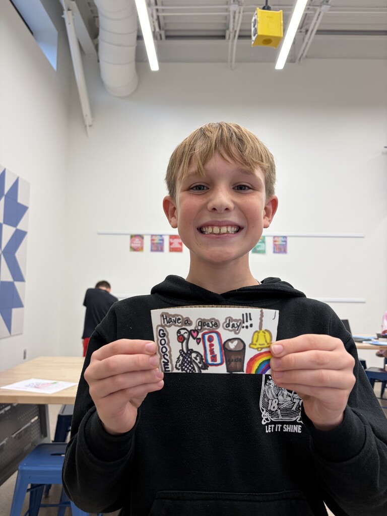 Smiling boy in a black hoodie holding a colorful handmade card with drawings and the message 'Have a great day!!' inside a classroom.