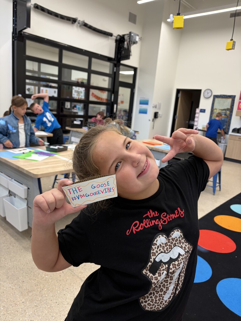 Smiling young girl in a Rolling Stones t-shirt holding a colorful handwritten sign that says '#VMGOOSEVIBES' in a classroom setting.