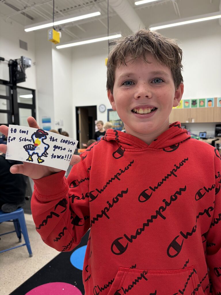 Smiling boy in a red Champion hoodie holding a handmade sign with a colorful goose drawing and the text 'Too cool for school, the coolest goose in town!!!'