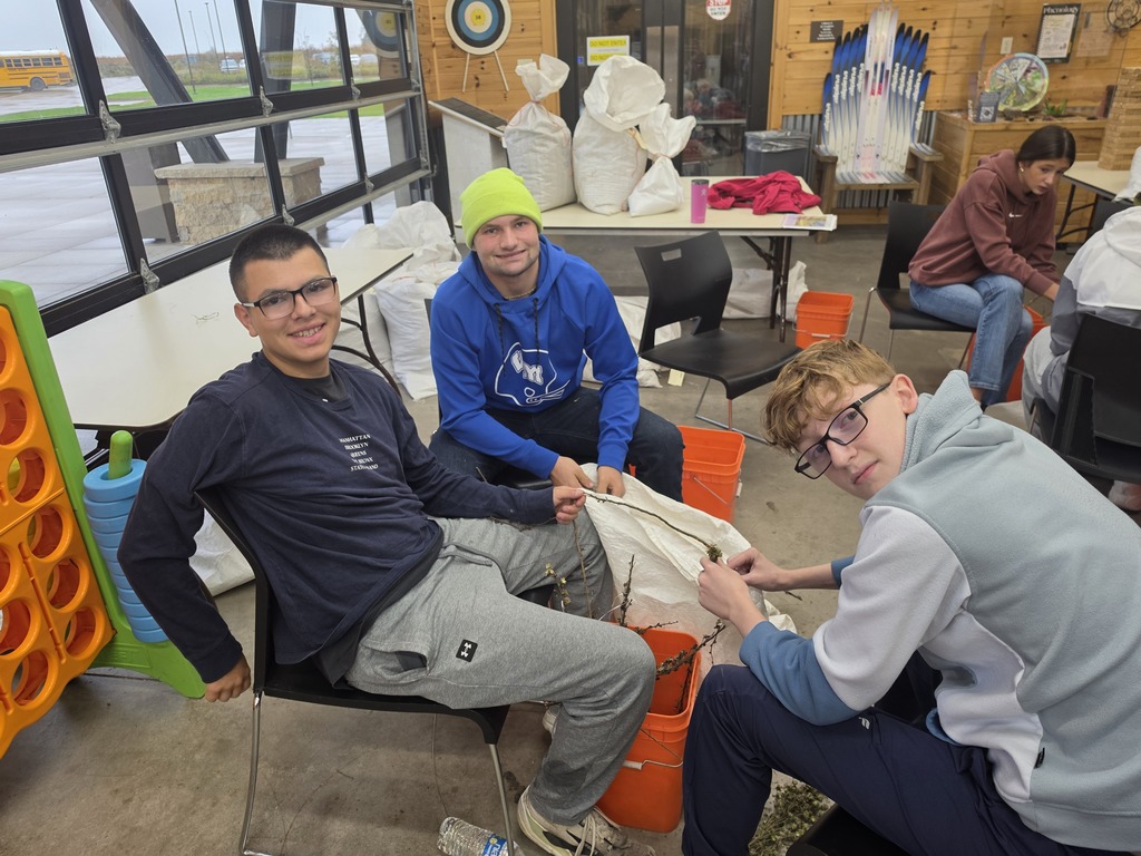 Three young men indoors sitting around a table, filling white sacks with materials, smiling at the camera in a casual setting.