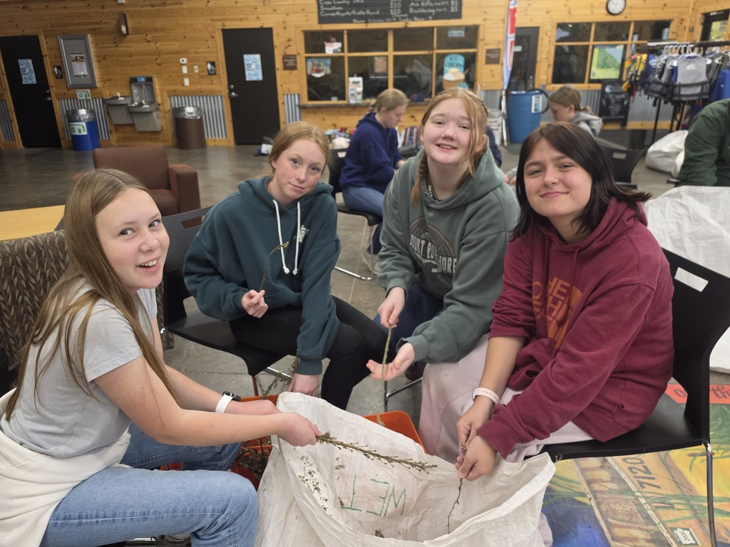 Four teenage girls sitting indoors, smiling and working together on a craft project involving a large white fabric piece.