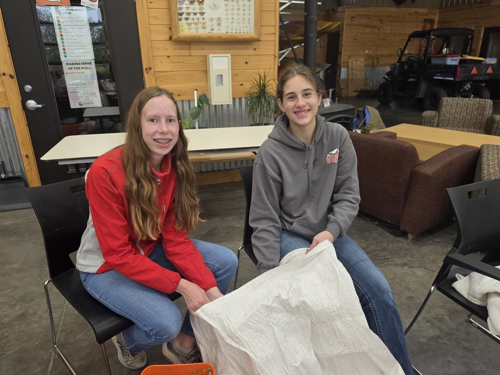 Two teenage girls sitting indoors on black chairs, smiling and holding a large white fabric or bag between them.