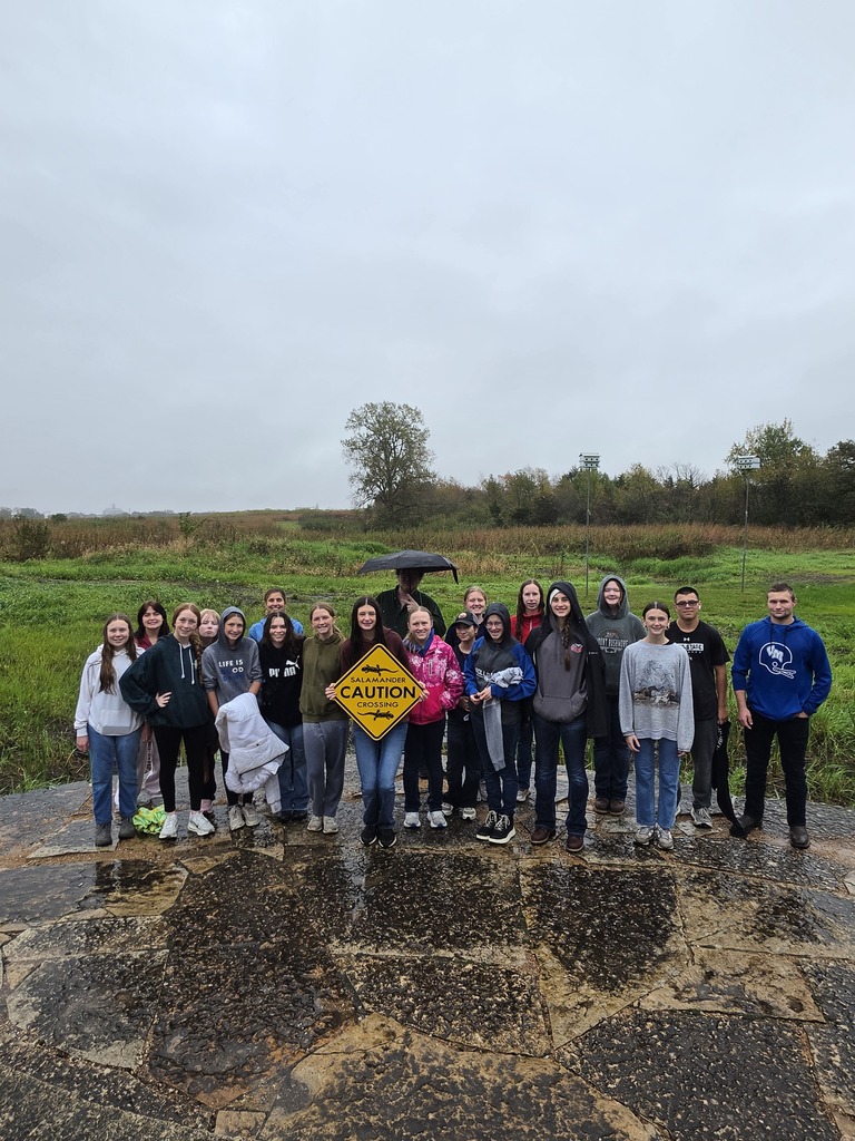 Group of young people standing outdoors on a wet stone surface with a grassy field and cloudy sky behind them.