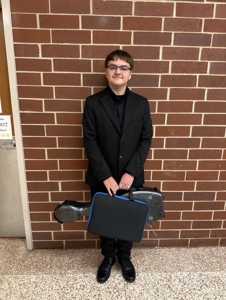 Student wearing a black suit and glasses stands in front of a brick wall, holding two cases.