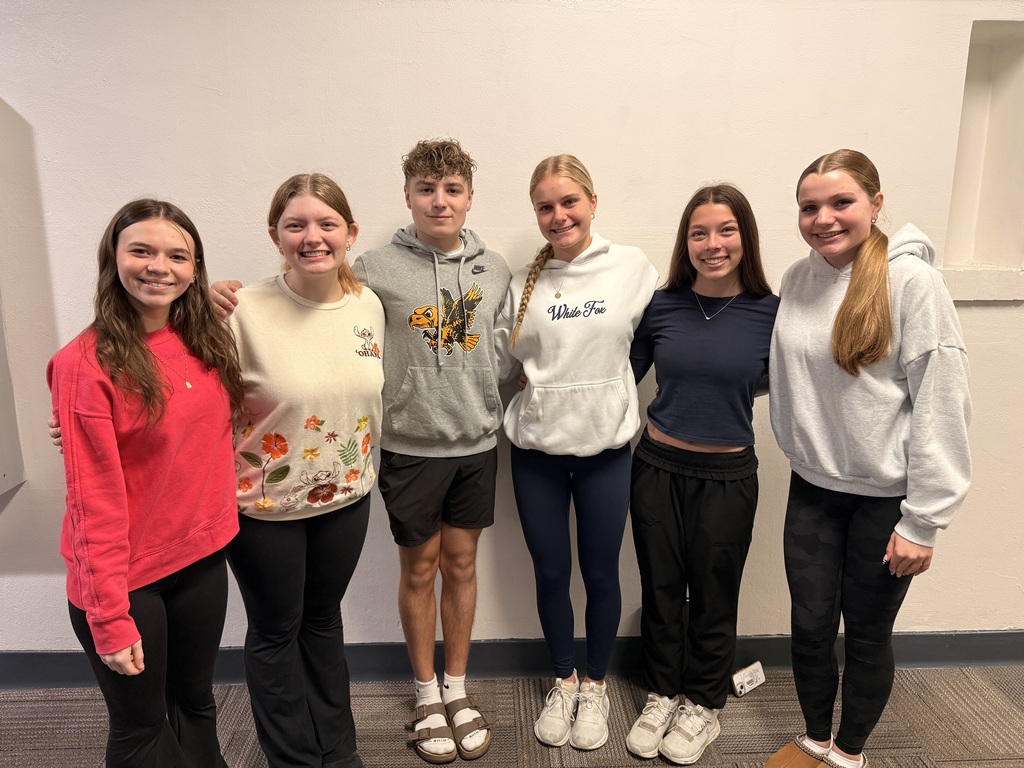 Six smiling high school students stand together with their arms around one another, posing against a white wall.