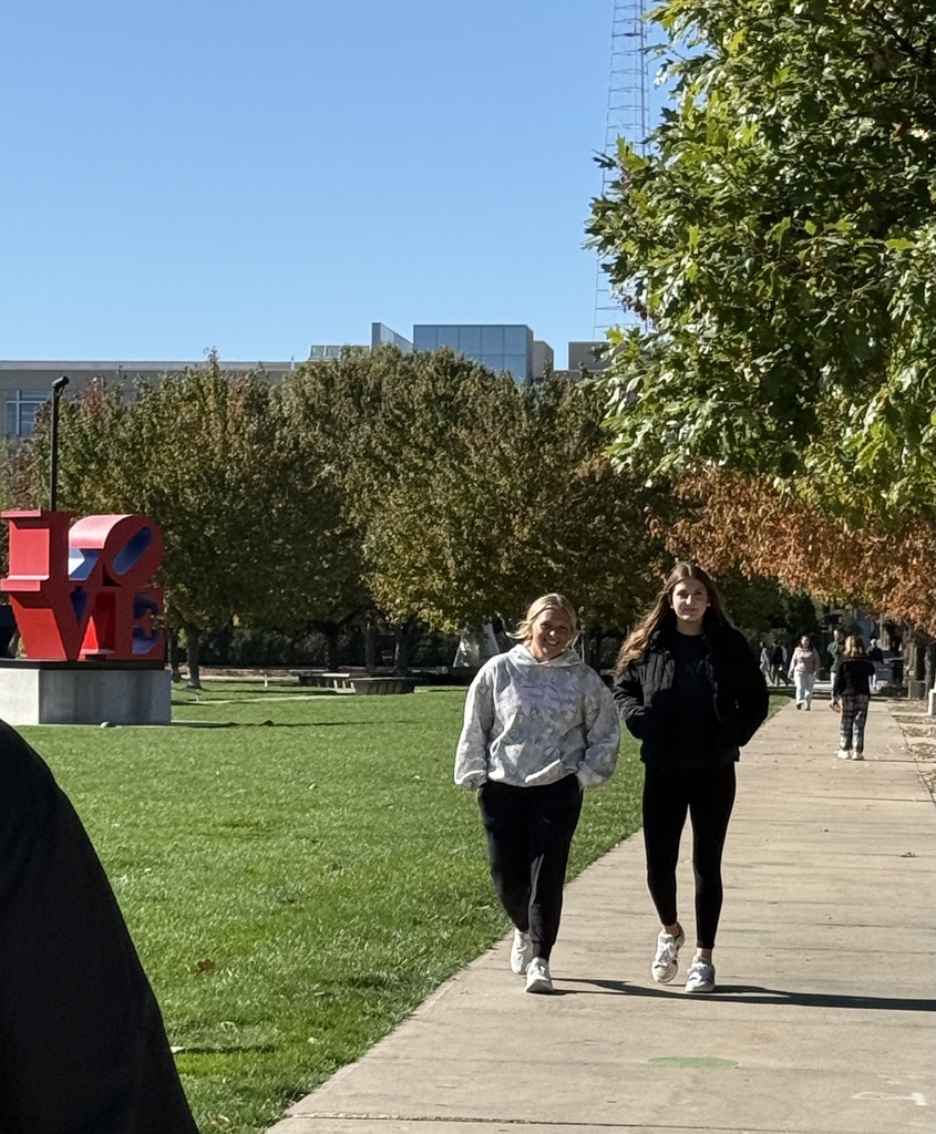 Two students walk along a sunny, tree-lined campus sidewalk near a large red LOVE sculpture.