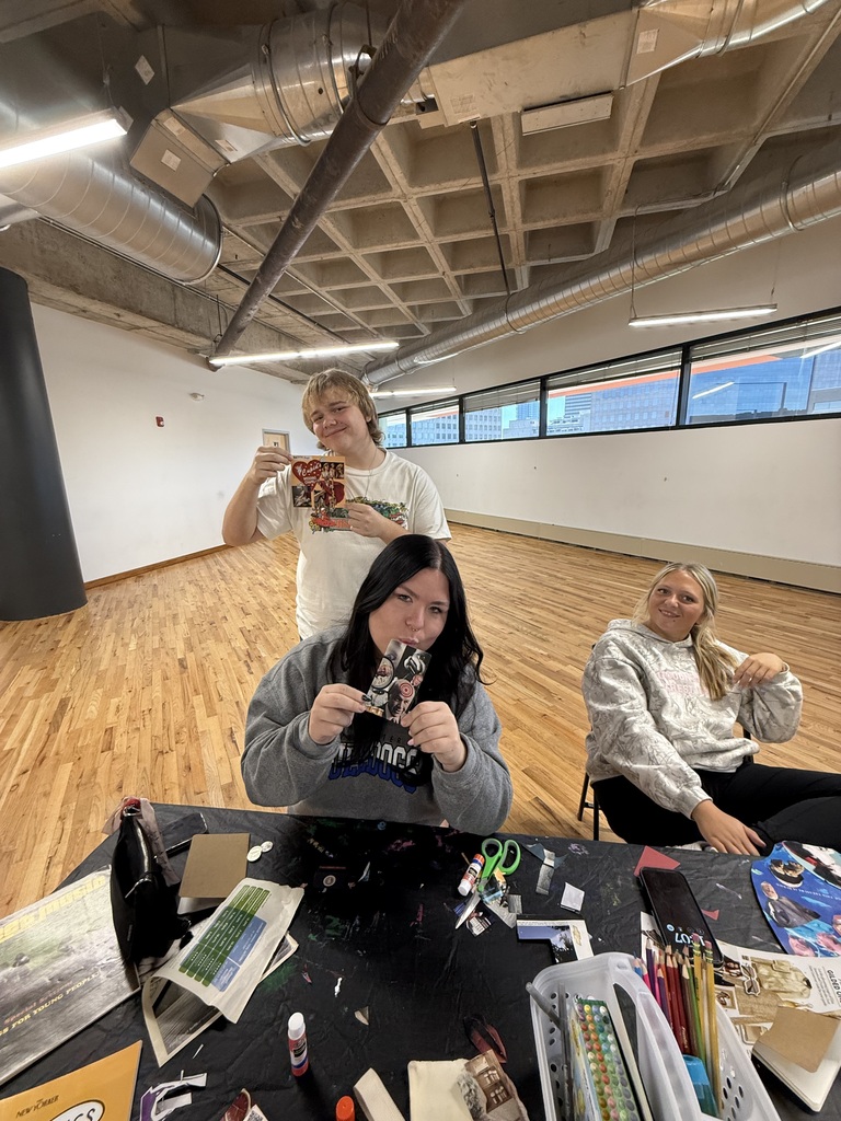 Three students proudly display their collage creations around an art table in a spacious studio with wooden floors.