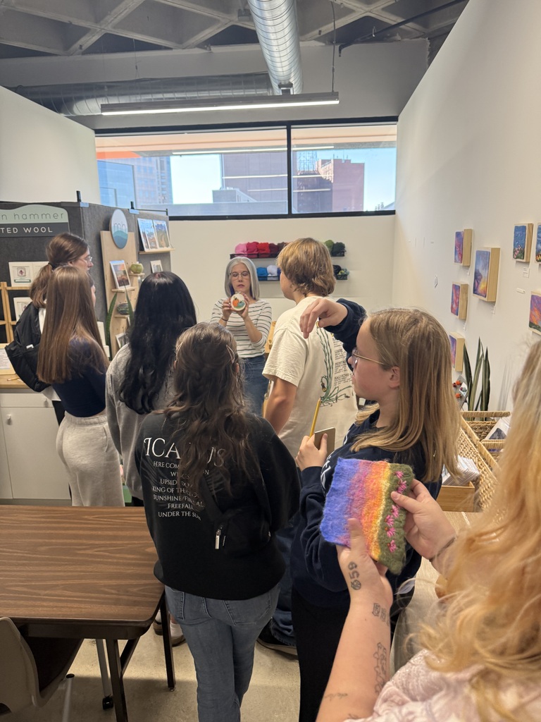 Students explore a colorful art gallery as a woman demonstrates a small handmade item surrounded by artwork and crafts.