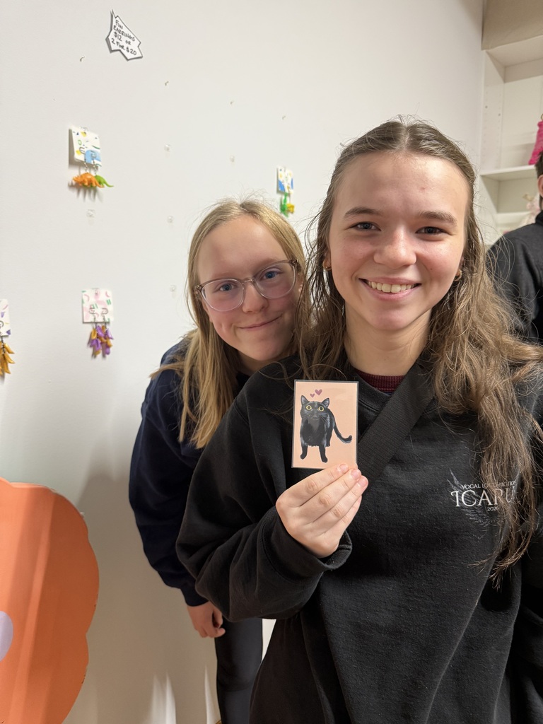 Two smiling students pose in an art gallery, one holding a small illustration of a black cat with a pink heart.