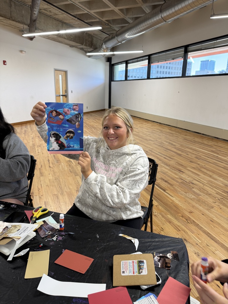 A smiling student in a gray hoodie holds up a colorful blue collage featuring people and animals during an art workshop.