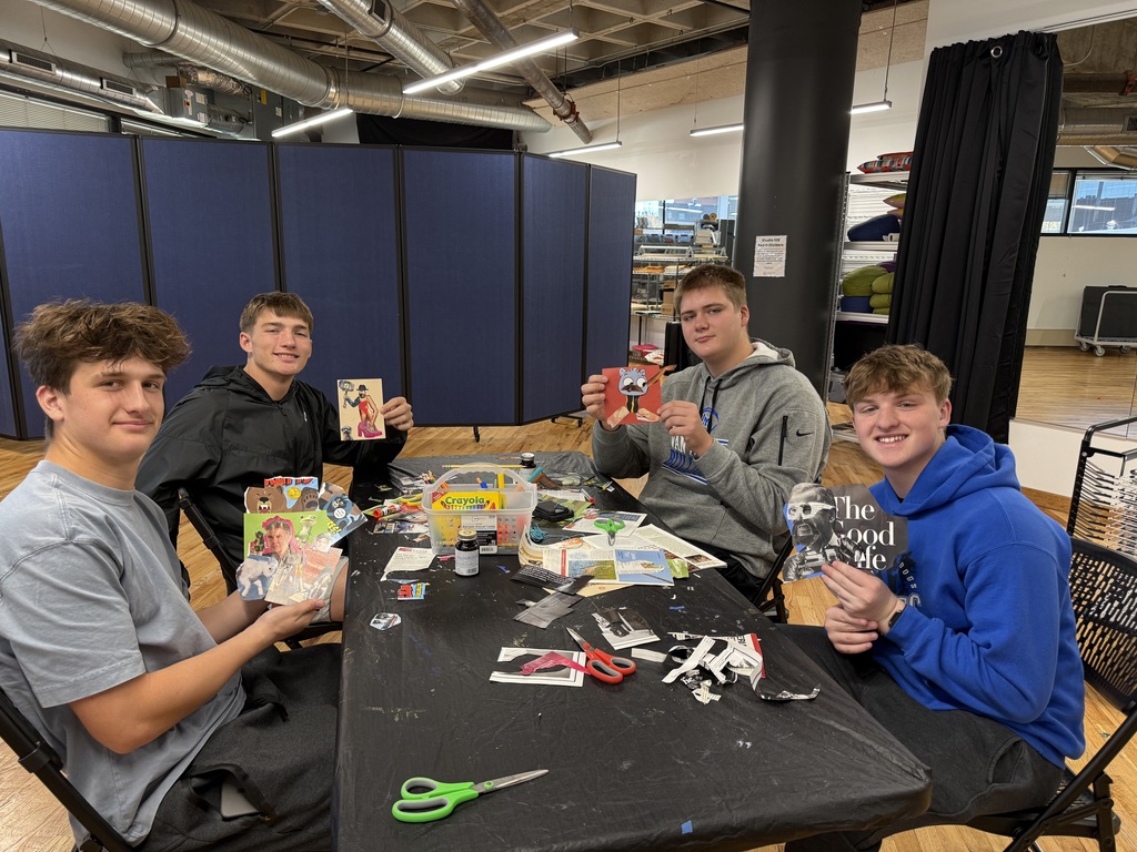 Four smiling students display handmade collages while seated around a table covered with art supplies and paper scraps.