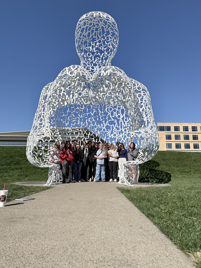 A group of students poses beneath a towering white sculpture of a seated human figure formed from interwoven letters.