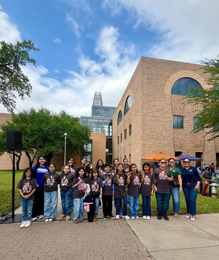 A group of people in matching shirts stands in front of a building. Trees and grassy areas surround them.