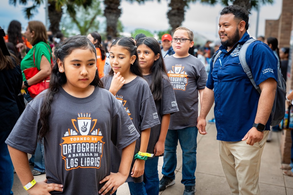 Four girls and a man, wearing matching gray t-shirts, stand on a sidewalk with others. The man wears a blue shirt and has a backpack.