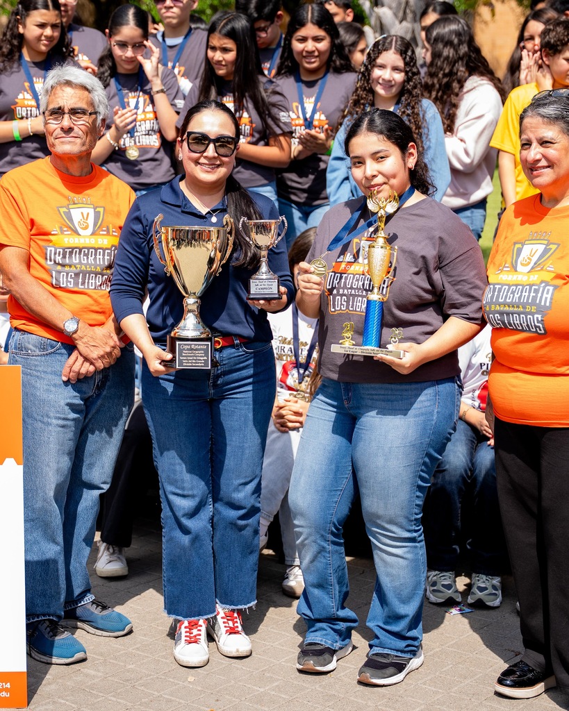A group of people pose with trophies in a schoolyard. Some wear casual clothes and sunglasses.