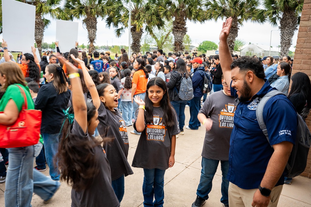 A group of people, including children and adults, in a crowded outdoor area with trees, raising hands.