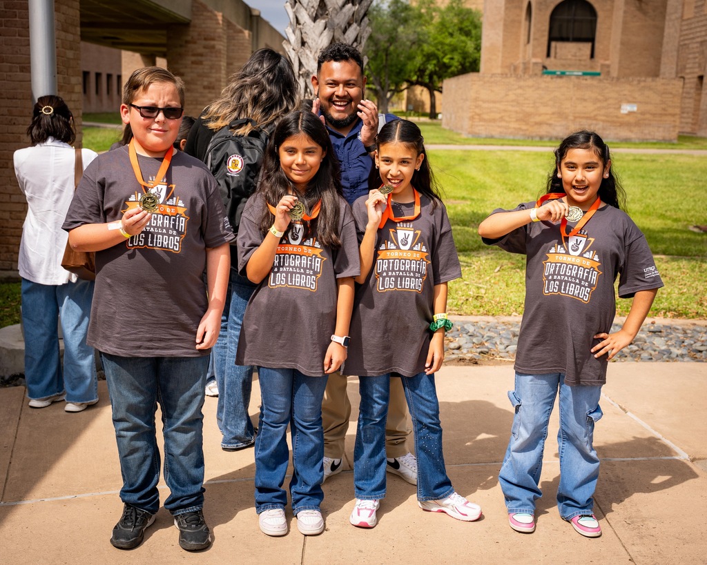 A group of children in matching shirts with medals pose on a sidewalk. Behind them is a building and people.
