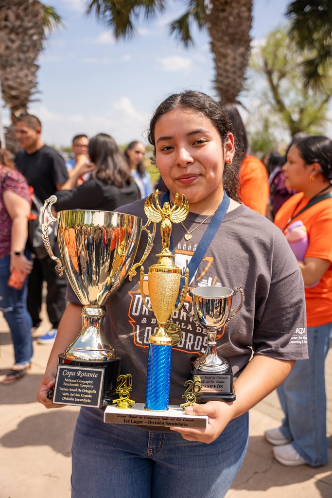 Woman in jeans and a gray shirt displays multiple trophies in front of a crowd.