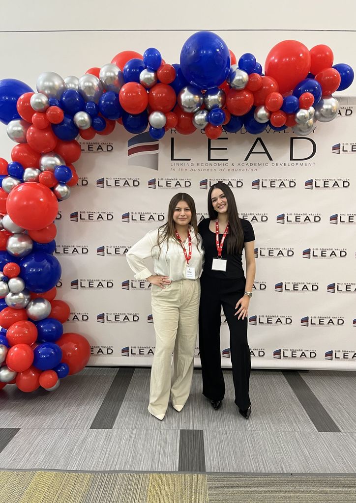 Two women stand in front of a large arch of red, blue, and silver balloons with "LEAD" on a white wall.