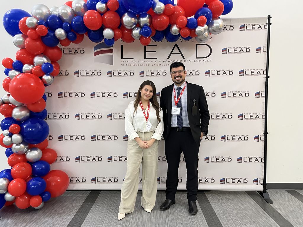Two people stand in front of a large red, blue, and silver balloon arch that says "LEAD" with a banner behind them.