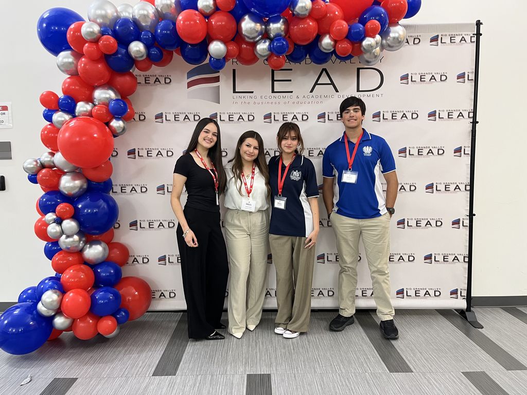 Four people stand together in front of a banner reading "LEAD". Behind them, a large balloon arch of red, white, and blue balloons.