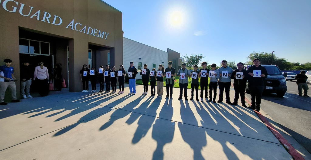 Group of people stand in line holding signs outside Guard Academy, shadows visible on ground.