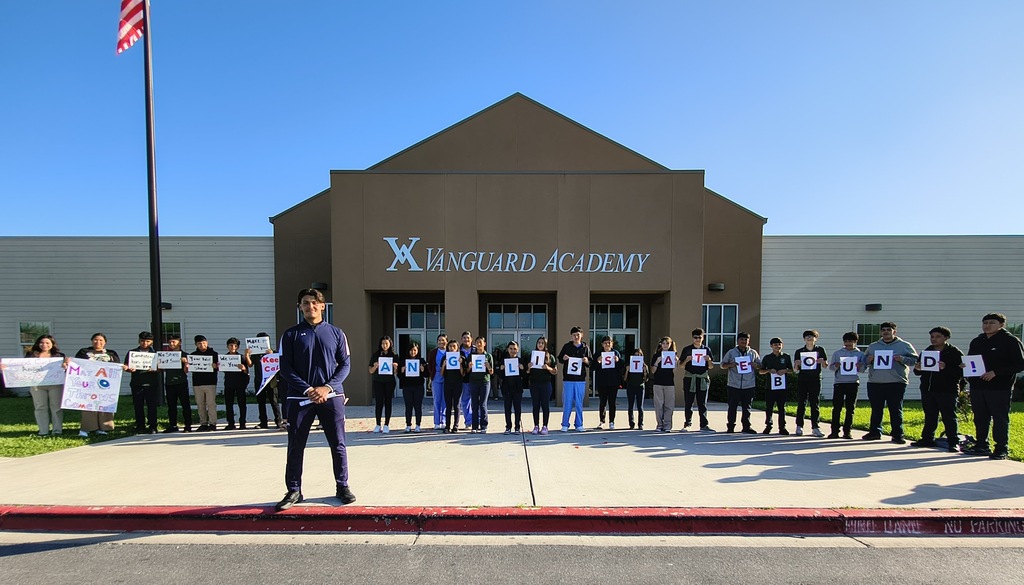 A man stands in front of a building with a sign reading "Xungard Academy." Behind him, students hold signs.