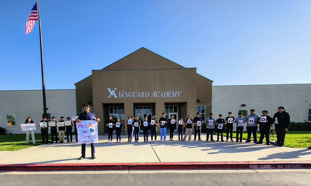 A group of individuals stands in front of a building. They hold signs. One person stands slightly forward.