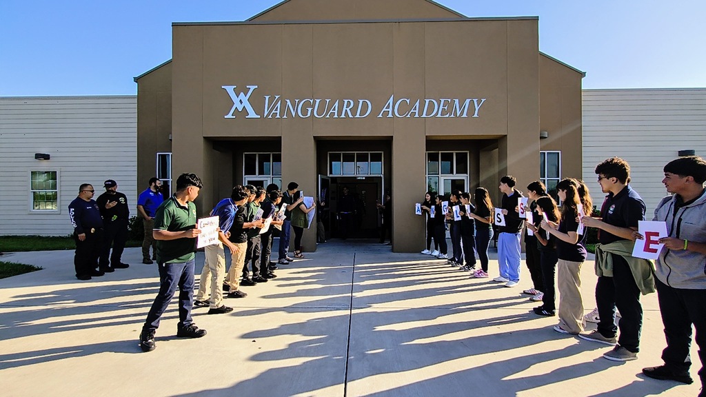 A line of students holding signs stands in front of Vanguard Academy. The building is brown and white.