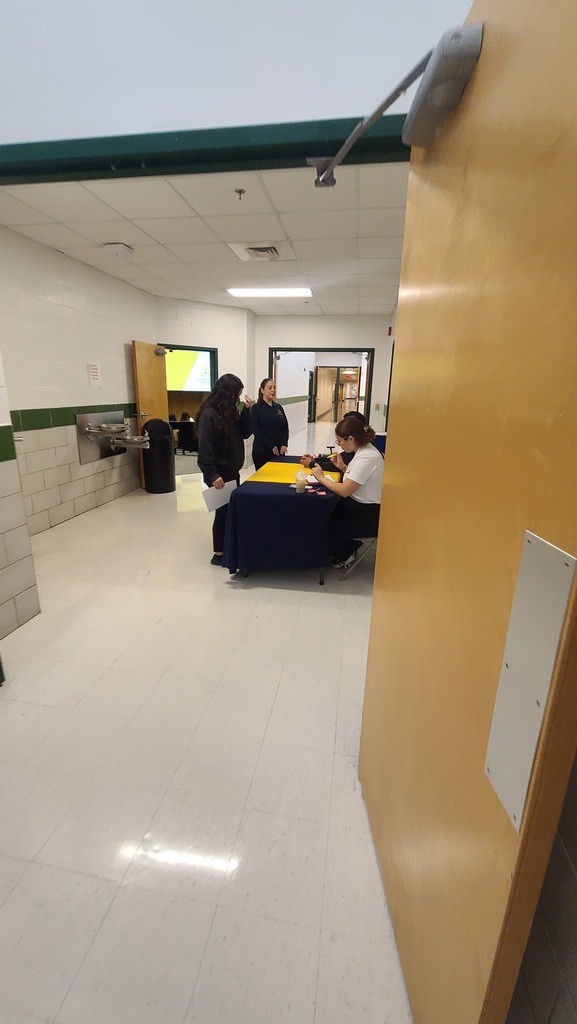 Three people are gathered around a table in a school hallway, with a yellow tablecloth and a trash bin.